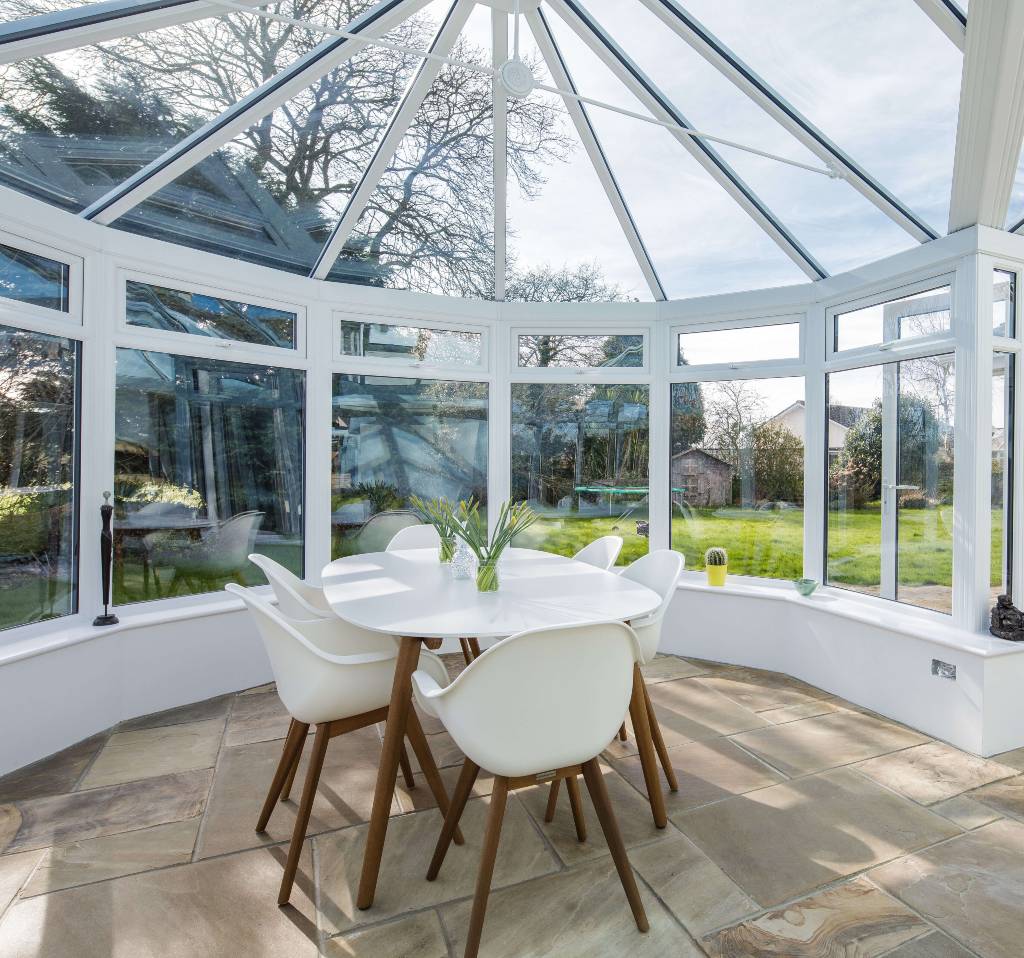 Interior of a modern conservatory with a round dining table and white chairs, offering a panoramic view of a lush garden through large glass windows and a glass roof.
