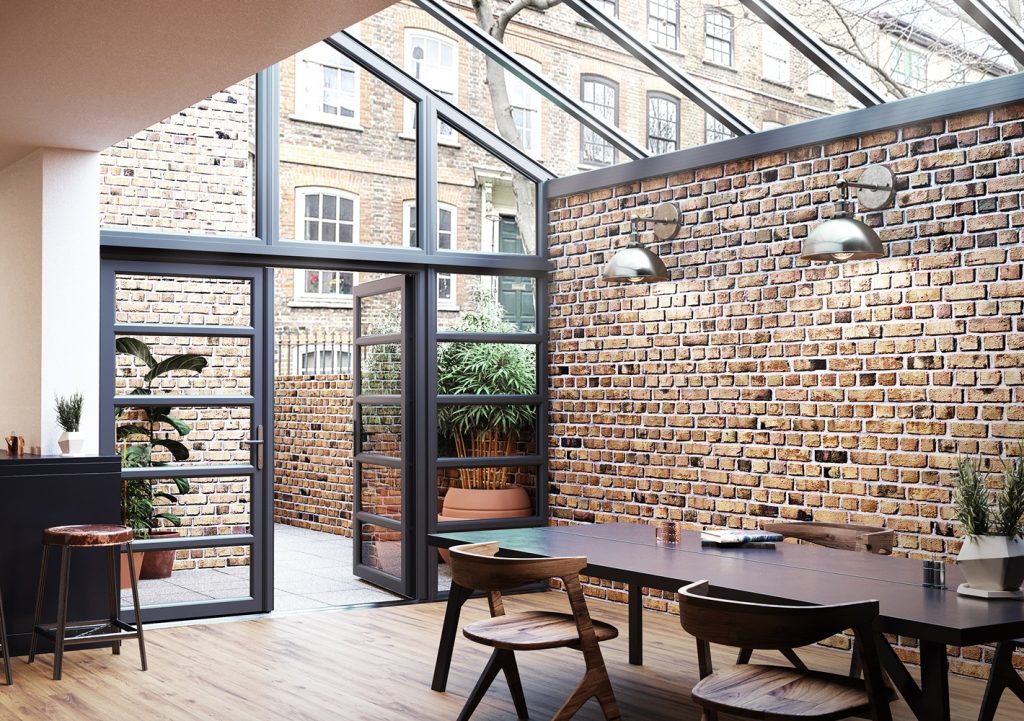 Interior of a modern conservatory with large glass panels and exposed brick walls, featuring stylish furniture and a view of an outdoor area through glass doors.